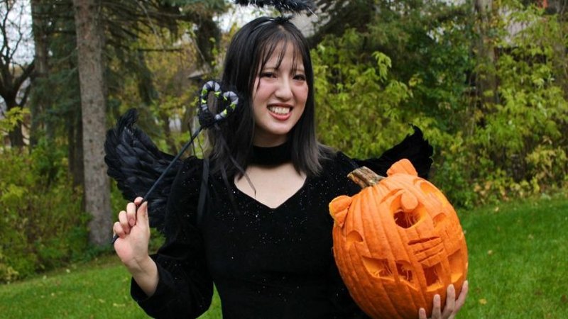 A person dressed in a black costume with black feathered wings and a halo headband is holding a carved pumpkin with a cat face design.