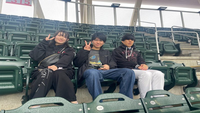 Three students sitting in the Twins stadium at a baseball game, posing with peace signs.