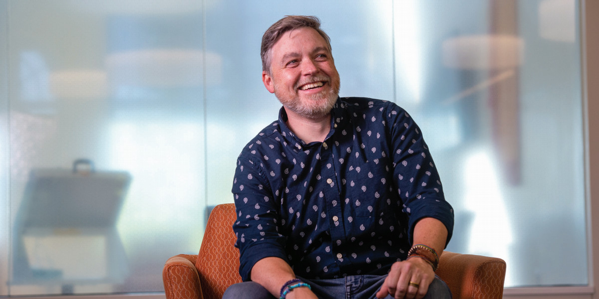 A man wearing a blue shirt sits in a chair and smiles for a photo