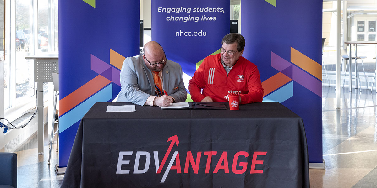 Two men sign a document on a table with the word Edvantage across the front